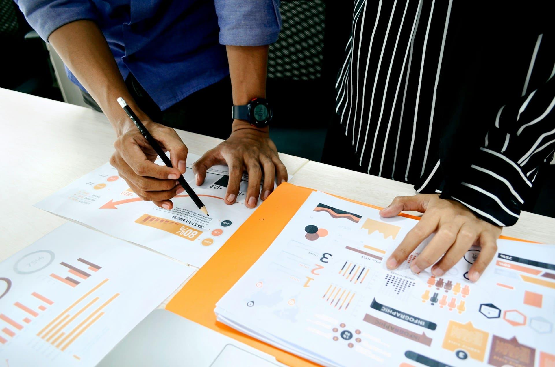 Two individuals collaborating on a project, surrounded by papers and pens, focused on their work.