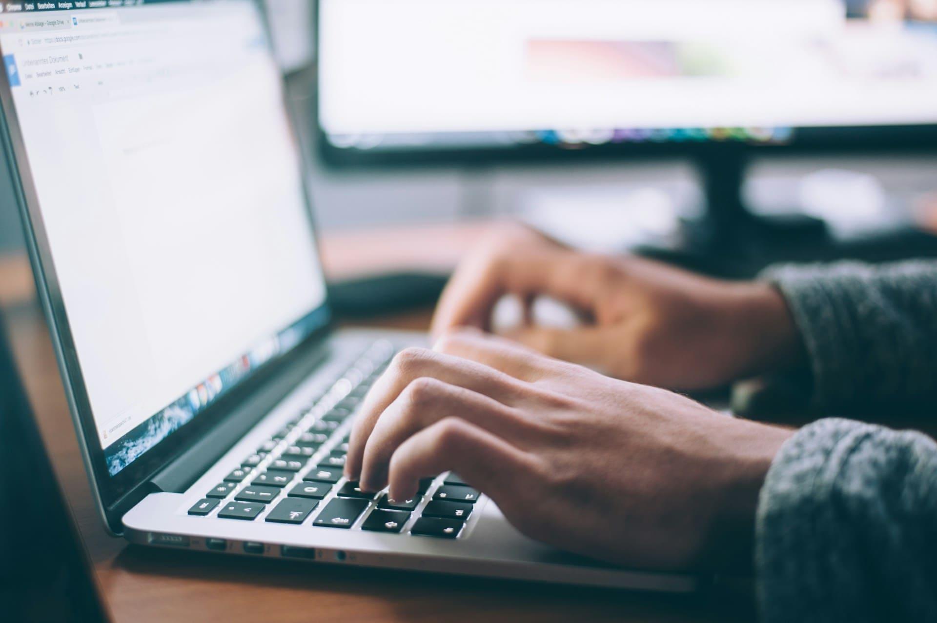 A person focused on typing on a laptop computer, with hands positioned on the keyboard and a screen visible in the background.