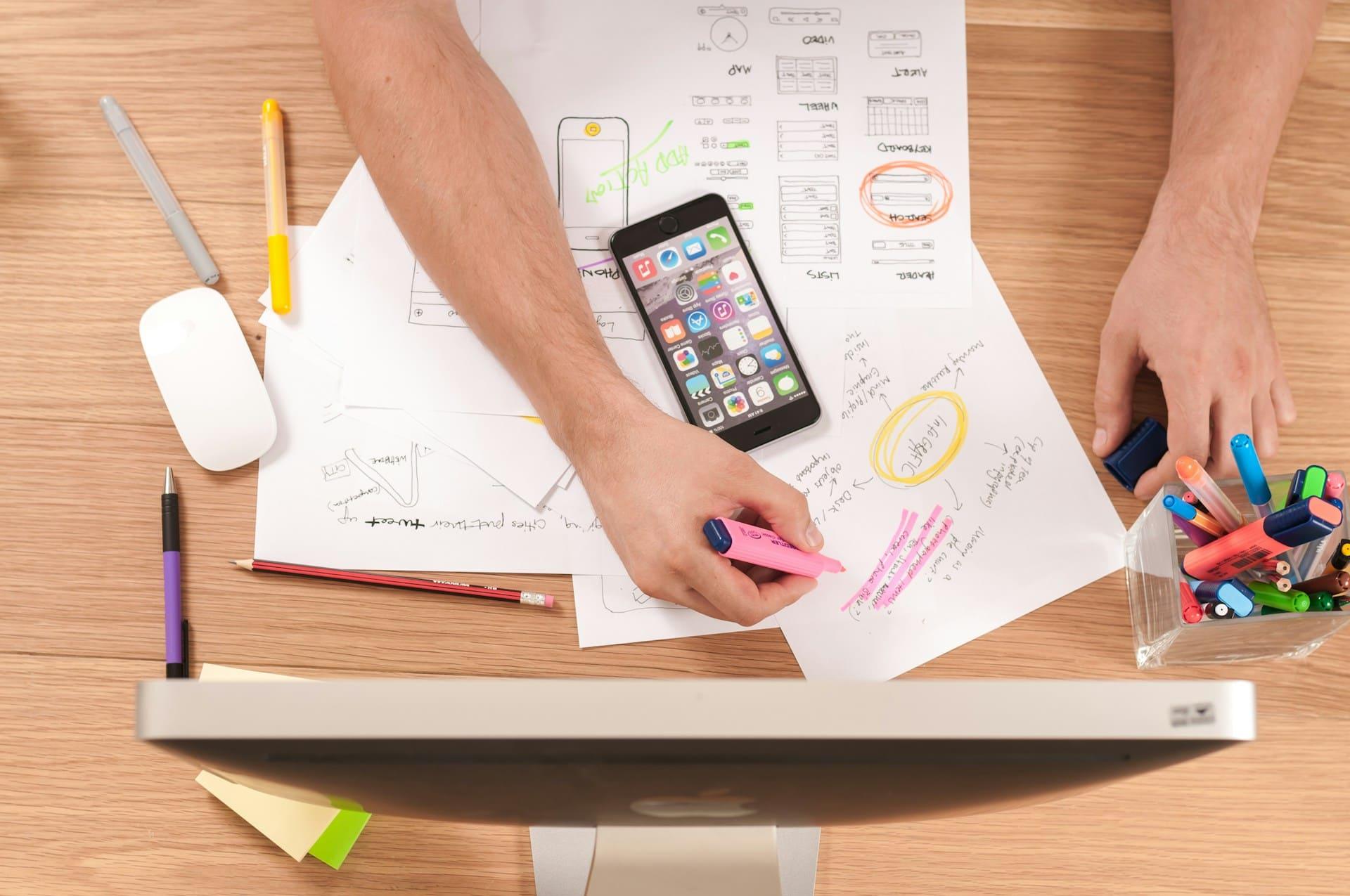 A person sitting at a desk, focused on using a cell phone with various office supplies in the background.