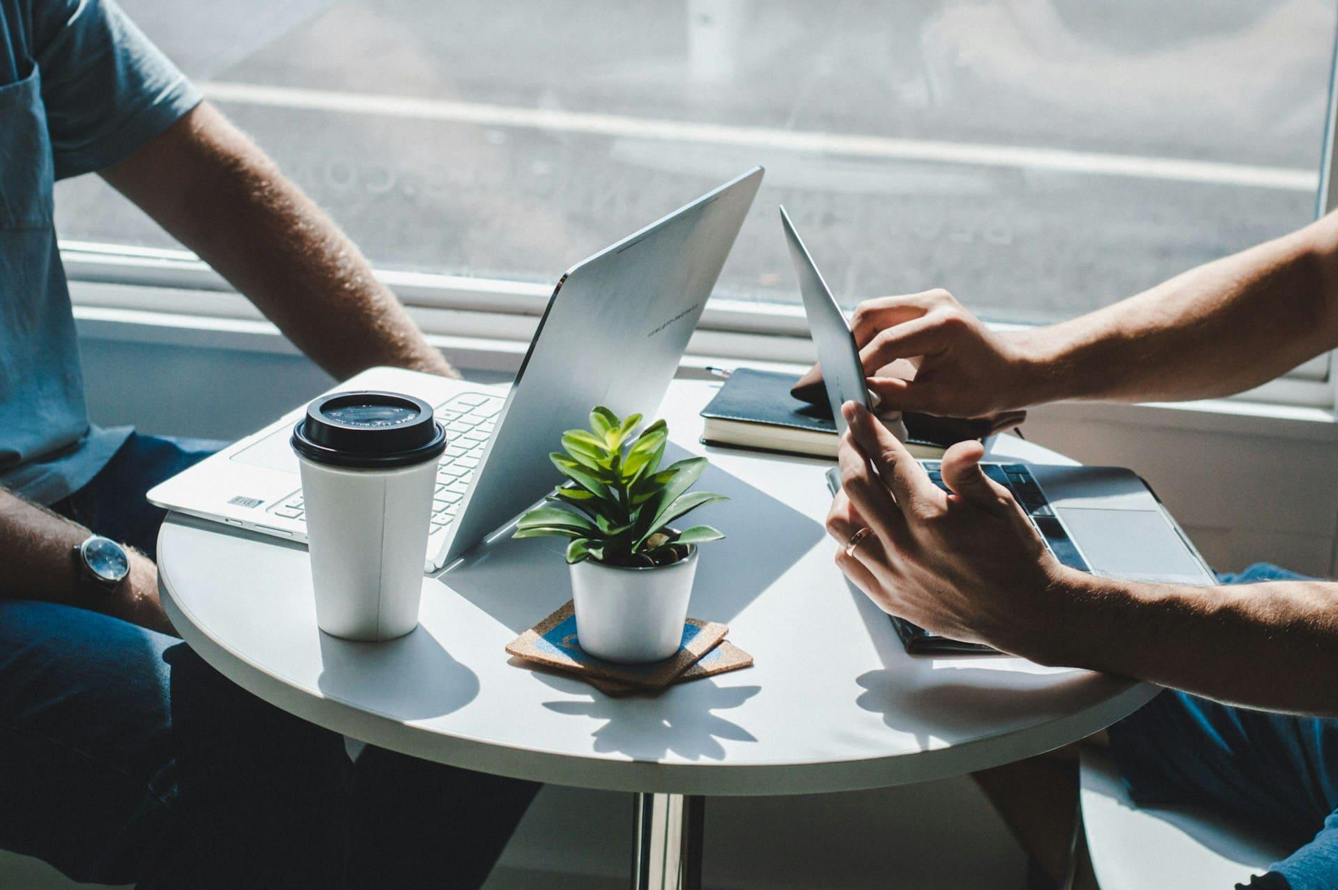 Two individuals seated at a table, each using a laptop, engaged in a collaborative work session.