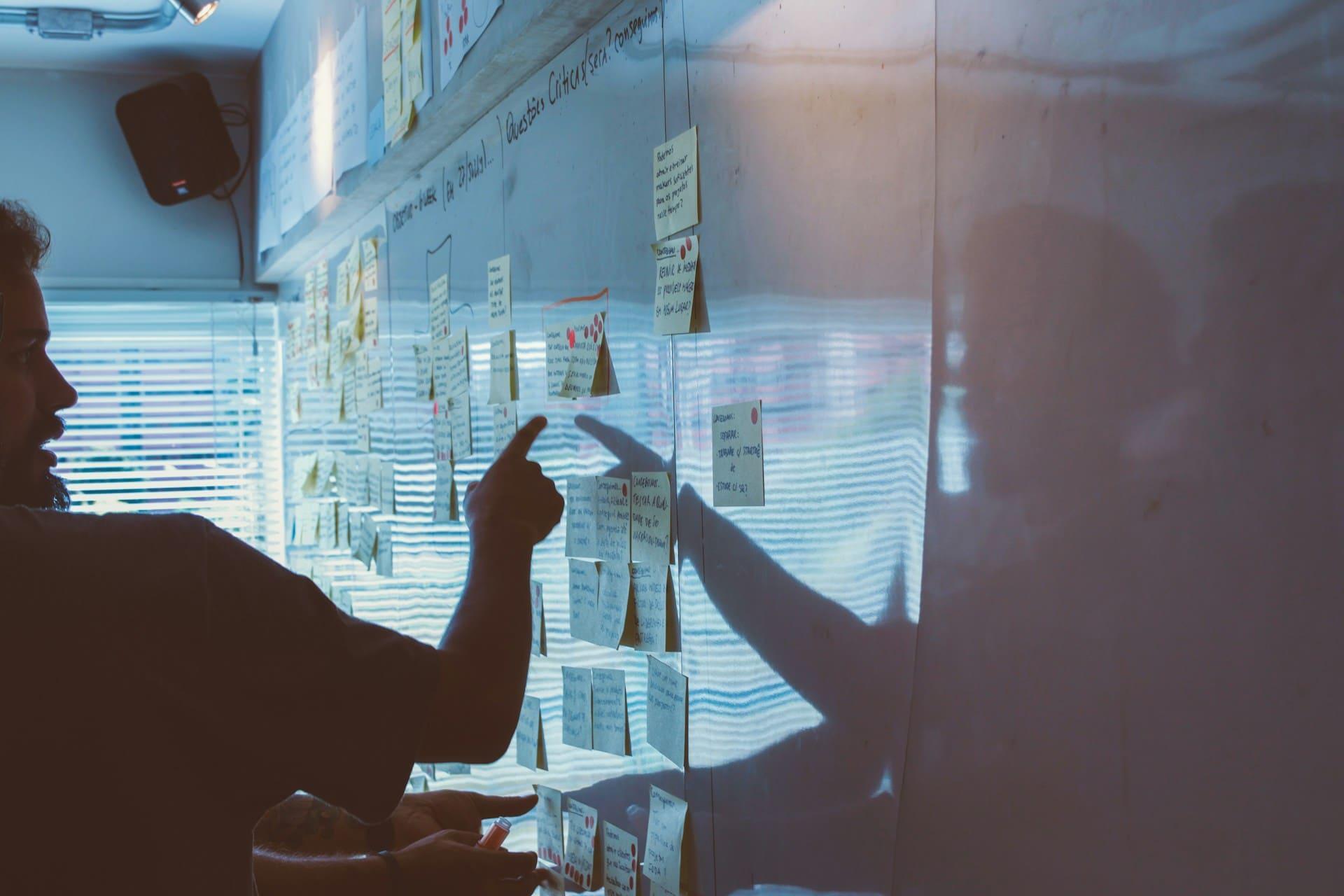 A man points at a whiteboard covered in colorful sticky notes, indicating a brainstorming or planning session.